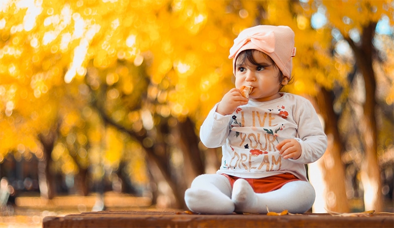 Baby sitting on table with fall leaves and trees behind her