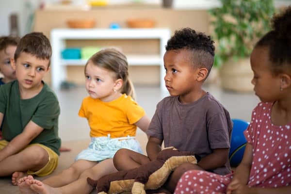Group of small nursery school children sitting on floor indoors in classroom, listening to teacher.